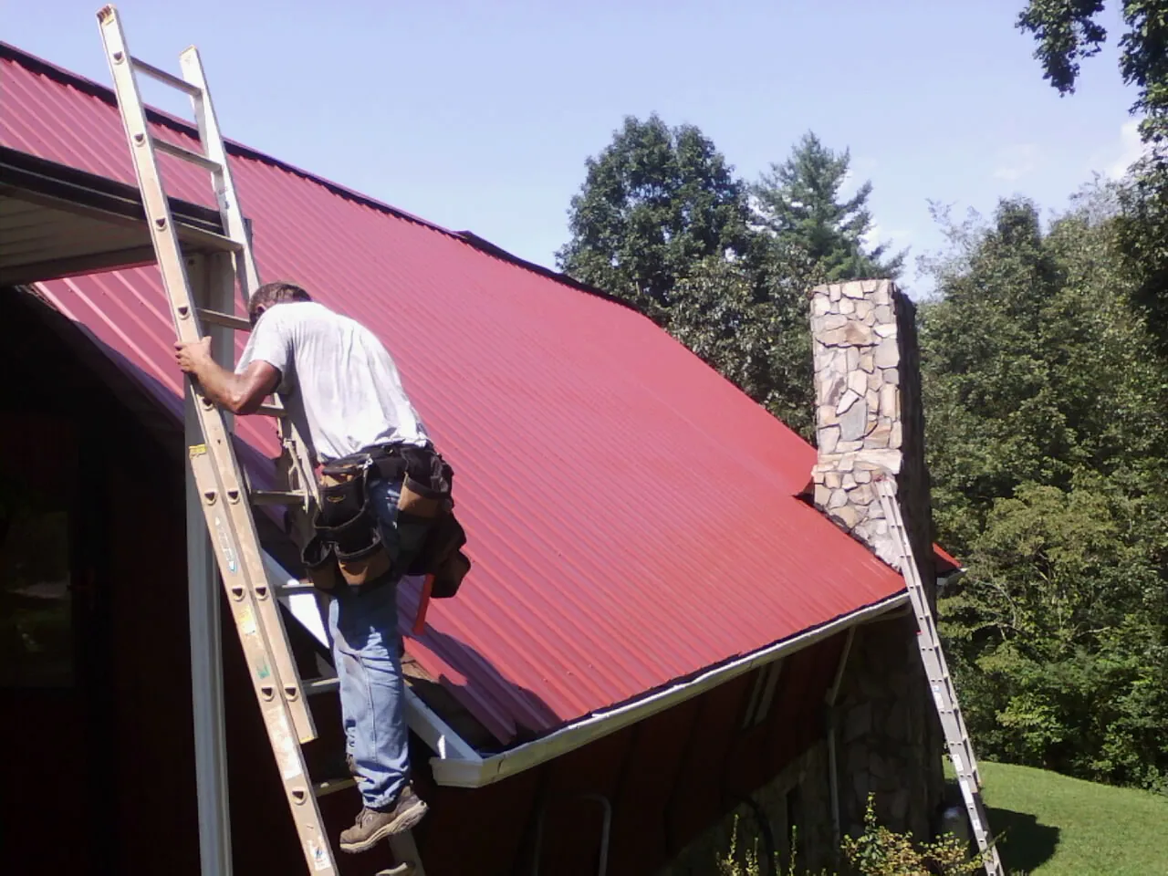 Roofer on a ladder beside a red metal roof and stone chimney — Soffit & Fascia Repair by Roofing Brothers in Lancaster